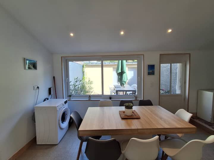 Salle à manger moderne avec table en bois, chaises blanches, lave-linge et portes ouvertes sur une terrasse ensoleillée