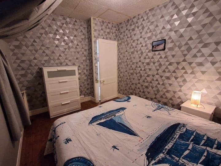 Petite chambre avec papier peint à motifs, commode blanche, lit, porte ouverte et lampe allumée sur la table de chevet.