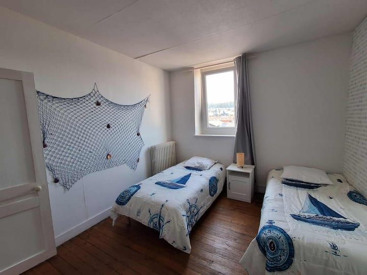 Petite chambre avec deux lits simples, linge de lit à thème marin, une fenêtre et des murs blancs.