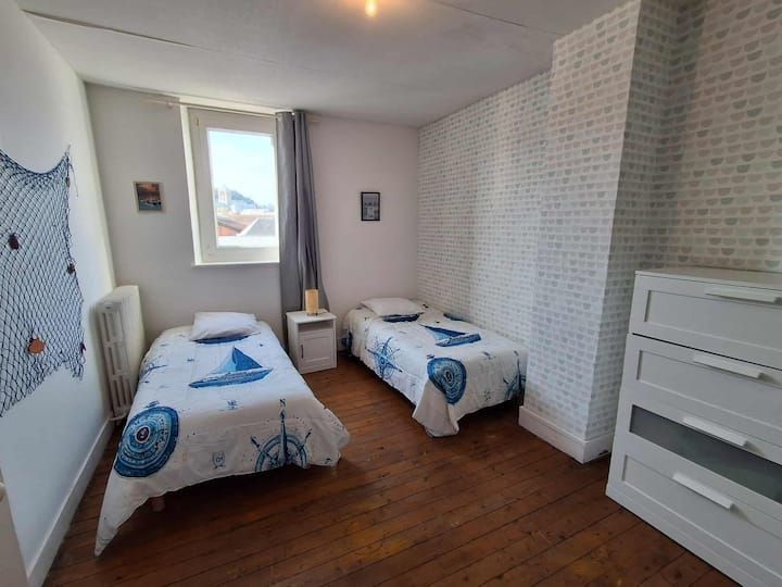 Petite chambre avec deux lits simples, murs blancs, parquet et une fenêtre avec des rideaux gris.