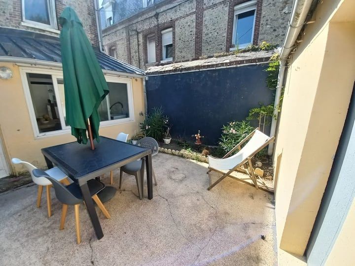 Petit patio avec une table, des chaises, un parasol vert fermé et une chaise longue adossée à un mur bleu.