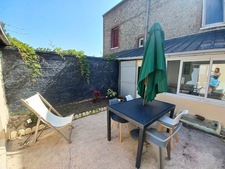 Terrasse avec table noire, parasol vert, chaise longue blanche et muret en pierre à côté d'une maison