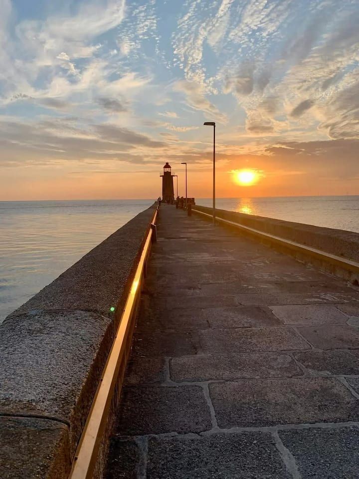 Coucher de soleil sur une longue jetée, une personne solitaire marchant vers un phare au bord de la mer
