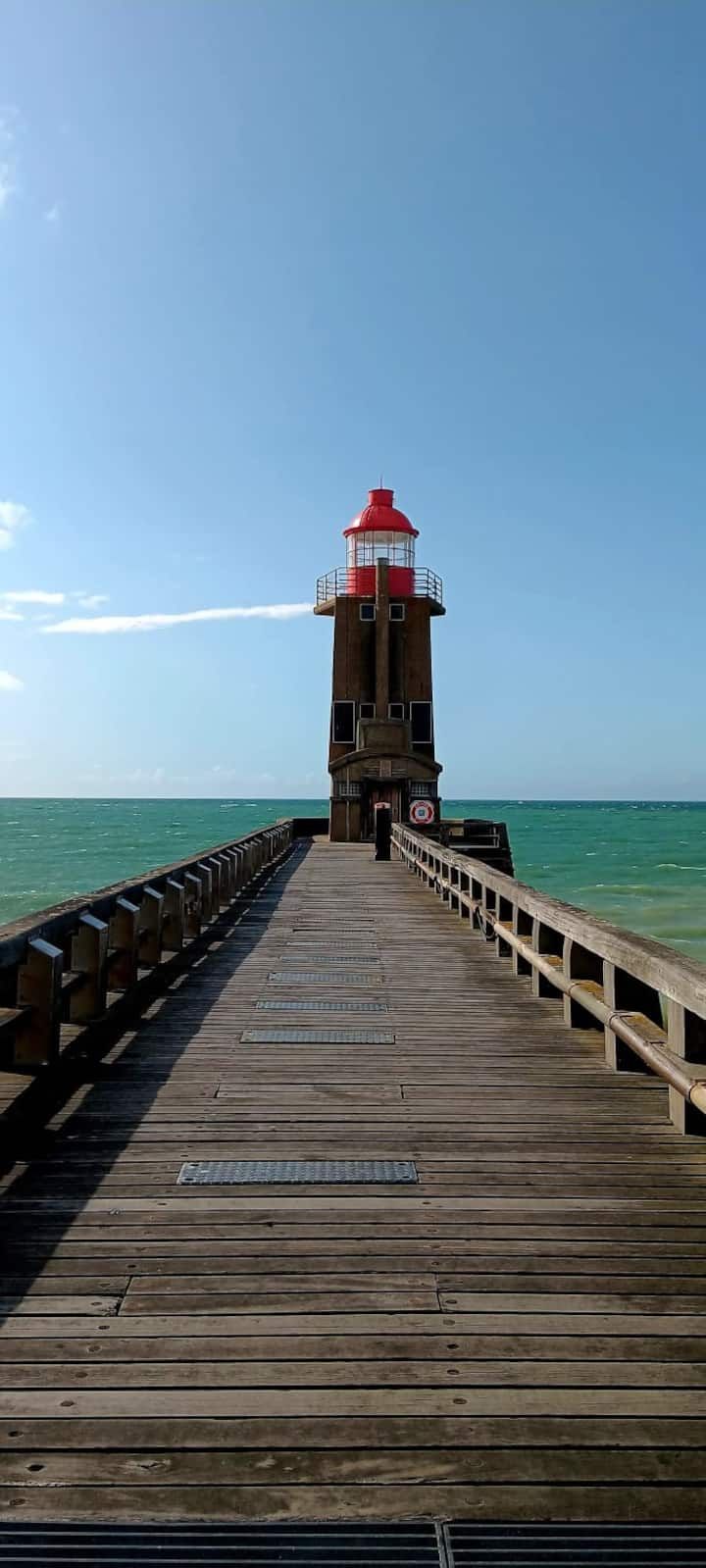 Jetée en bois menant à un phare rouge surplombant des eaux turquoise sous un ciel d'un bleu limpide