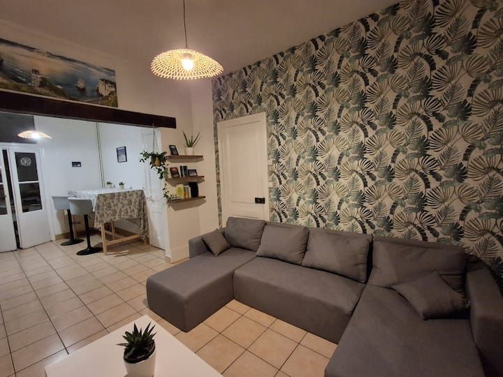 Salon moderne avec canapé d'angle gris, papier peint tropical, sol carrelé et coin cuisine ouvert