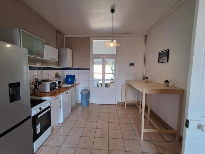 Cuisine étroite et lumineuse avec placards blancs, sol carrelé, réfrigérateur, four et table en bois près d'une porte.