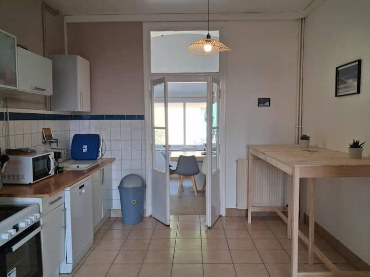 Petite cuisine avec placards blancs, sol carrelé, table en bois et porte donnant sur une salle à manger ensoleillée
