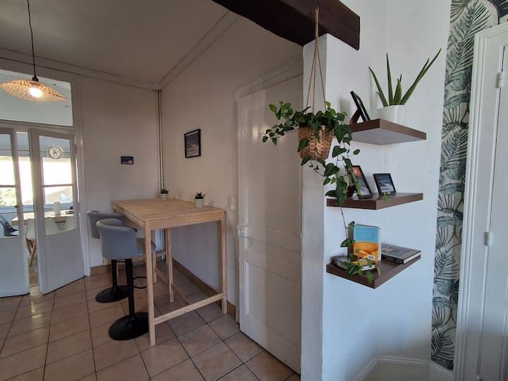 Petit coin cuisine avec une table en bois, des tabourets, un meuble blanc et des plantes suspendues, près d'un couloir carrelé.