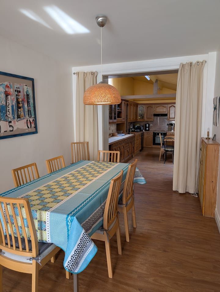 Salle à manger avec table en bois, chemin de table bleu et chaises donnant sur une cuisine chaleureuse.