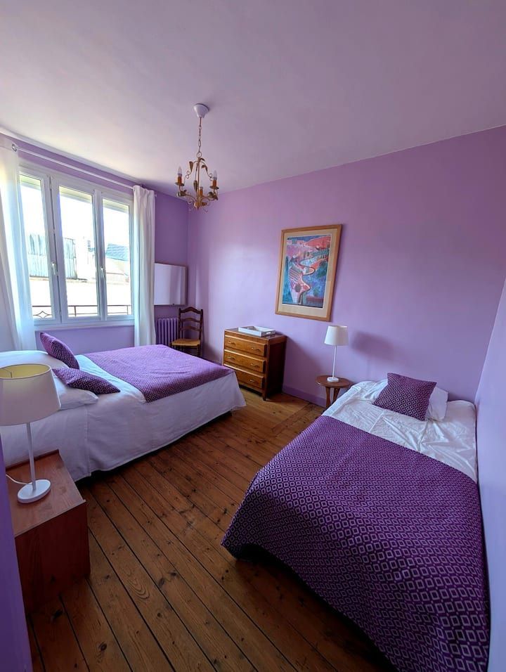 Chambre lavande lumineuse avec deux lits simples, parquet, commode, fenêtre et lustre.