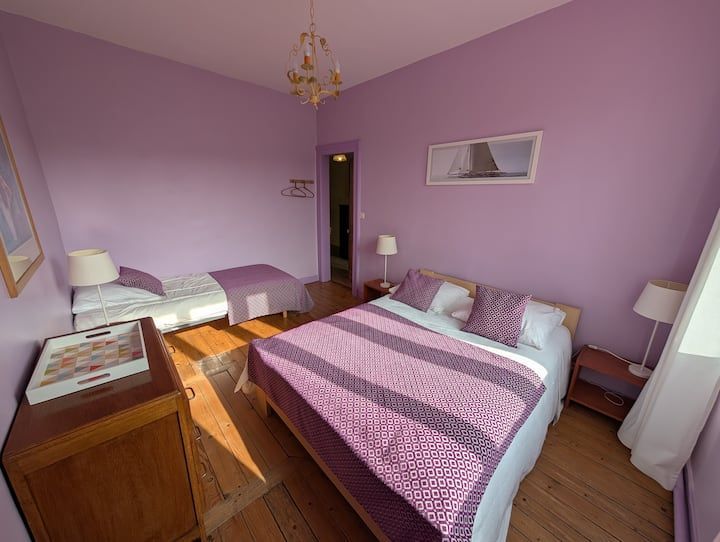 Chambre lavande avec deux lits, commode en bois et lumière du soleil entrant par la fenêtre.