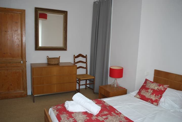 Une chambre modeste avec deux lits jumeaux, une commode en bois, un miroir, une chaise, une lampe rouge et du linge de lit.