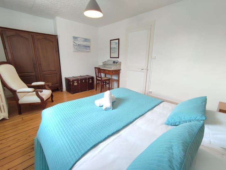 Chambre lumineuse avec couvre-lit turquoise, armoire en bois, bureau et porte blanche près du lit.