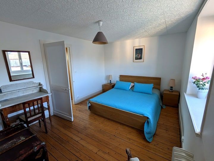 Petite chambre avec un lit, des draps bleus, un parquet, un bureau et une banquette près de la fenêtre, baignée de lumière naturelle.
