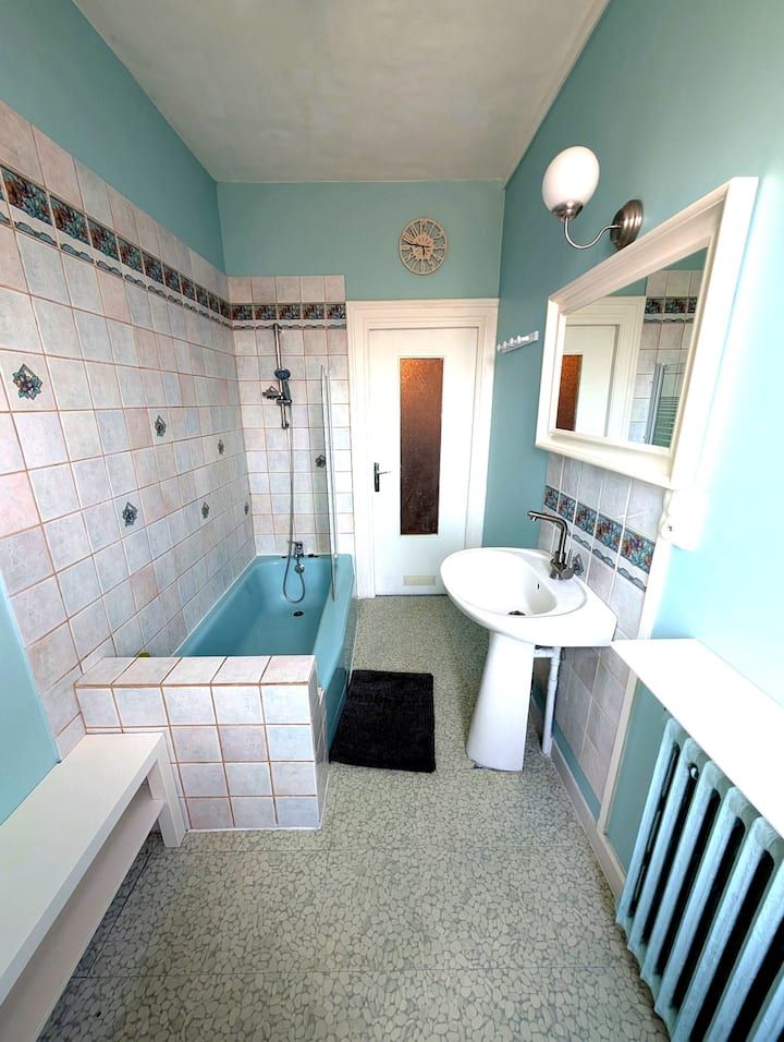 Petite salle de bains aux tons pastel avec baignoire carrelée de rose, lavabo blanc, miroir mural et fenêtre dépolie.