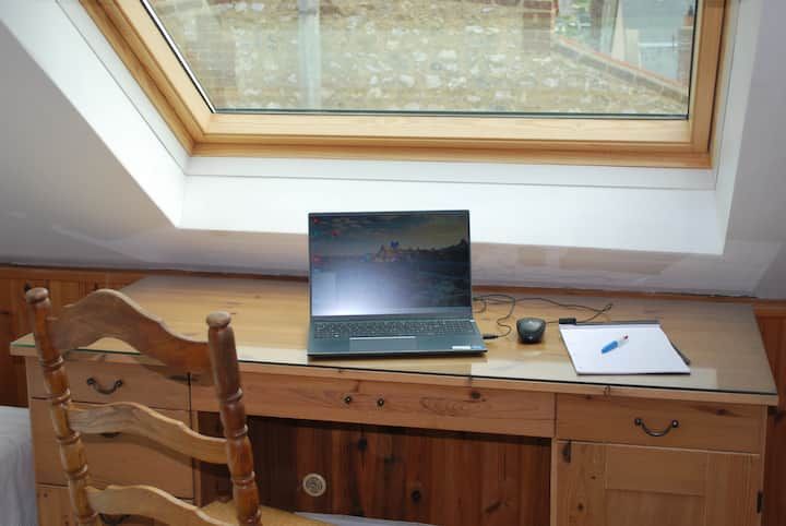 Un bureau en bois sous une verrière, avec un ordinateur portable ouvert, une souris et un carnet à côté d'une chaise.