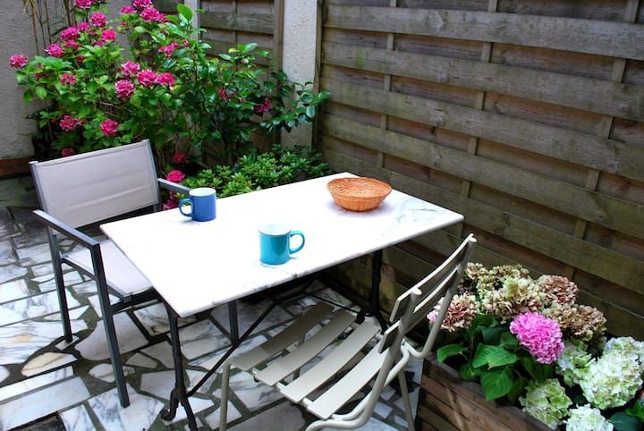 Petite table de patio avec des tasses et un bol à côté de chaises, entourée de fleurs et d'une clôture en bois.