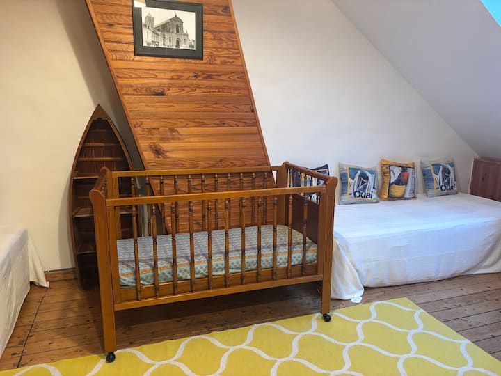 Chambre d'enfant avec un berceau en bois, un lit de jour blanc et un mur lambrissé incliné sous des tableaux encadrés.