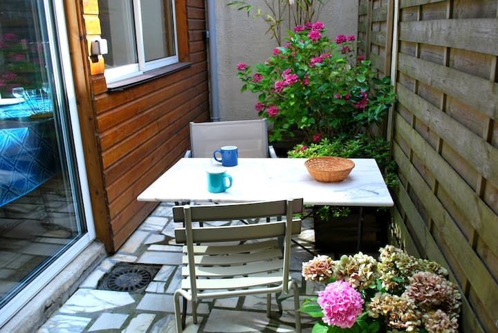 Petit patio avec table blanche, chaises, tasses et fleurs roses, à côté d'une clôture en bois et d'une porte coulissante.
