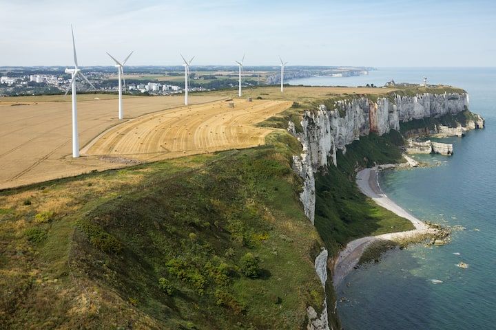 Éoliennes sur une falaise herbeuse surplombant une mer bleue, des falaises de craie blanche et un littoral lointain.