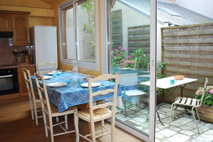 Coin repas de la cuisine donnant sur une terrasse avec nappe bleue, chaises blanches et fleurs en pot