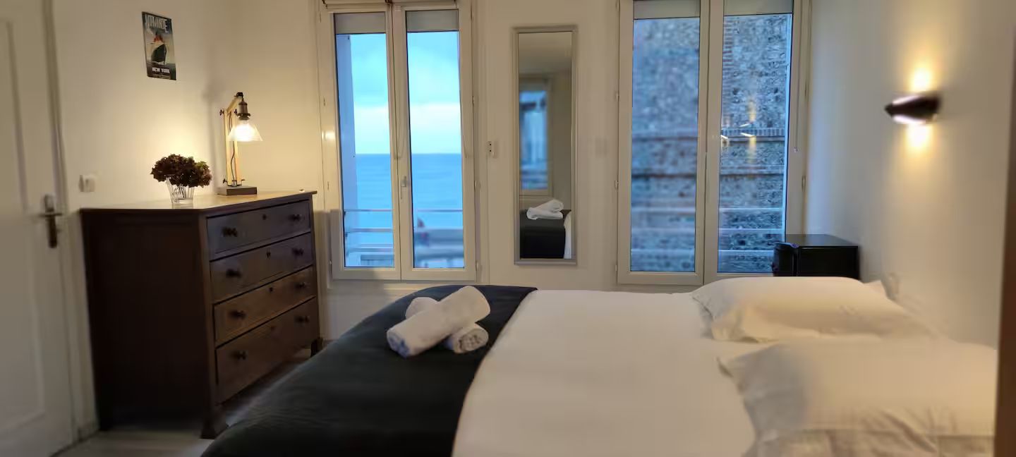 Une chambre avec un lit, une commode, un miroir et de grandes fenĂȘtres.