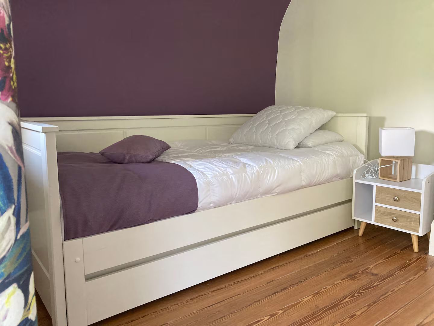 Une chambre avec un lit, une table de chevet, une lampe et un mur violet.