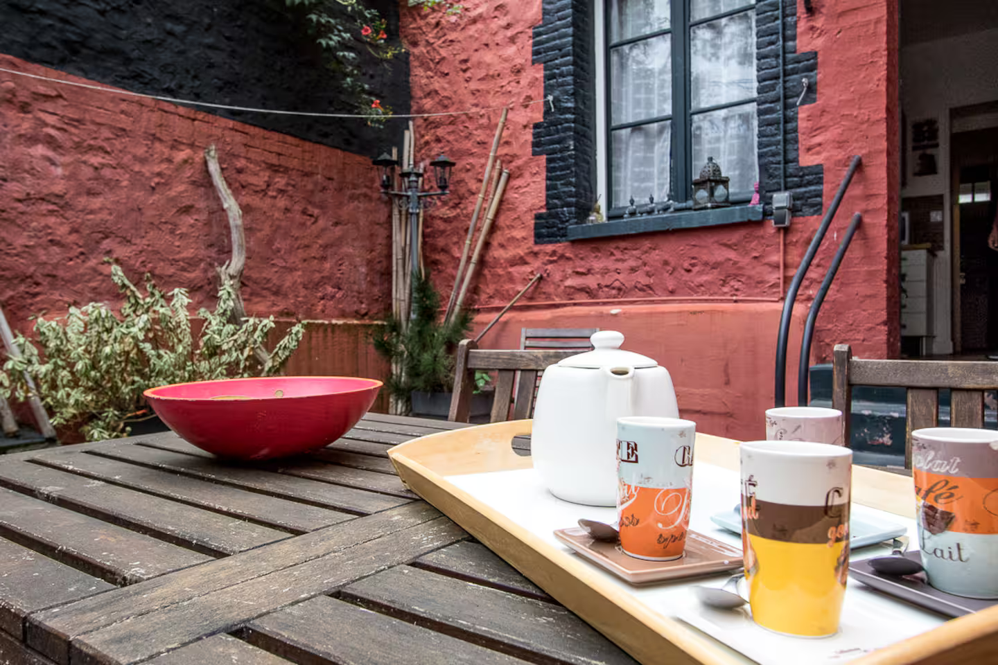 Une table en bois avec un bol et des tasses dessus devant un bâtiment rouge.
