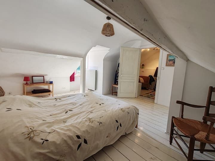 Chambre avec un lit, une chaise en bois, un sol blanc et une porte ouverte donnant sur une autre pièce.