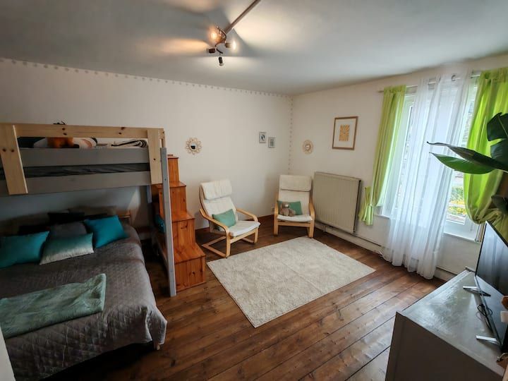 Chambre avec lit superposé, coin salon et fenêtre avec rideaux ; parquet et murs blancs.