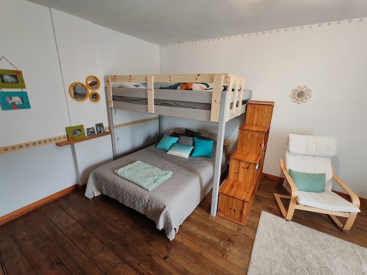 Chambre avec lits superposés. Le lit du bas est fait, un escalier en bois mène au lit du haut. Chaise blanche et tapis.