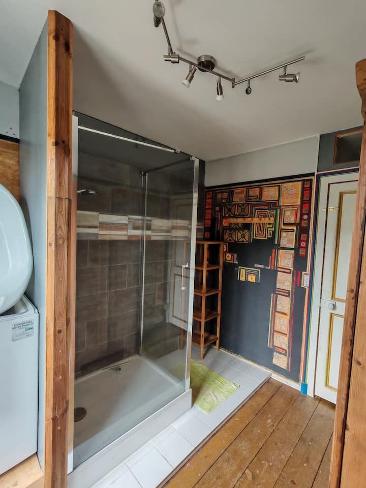 Salle de bains avec douche en verre, poutres en bois, machine à laver et parquet brut.