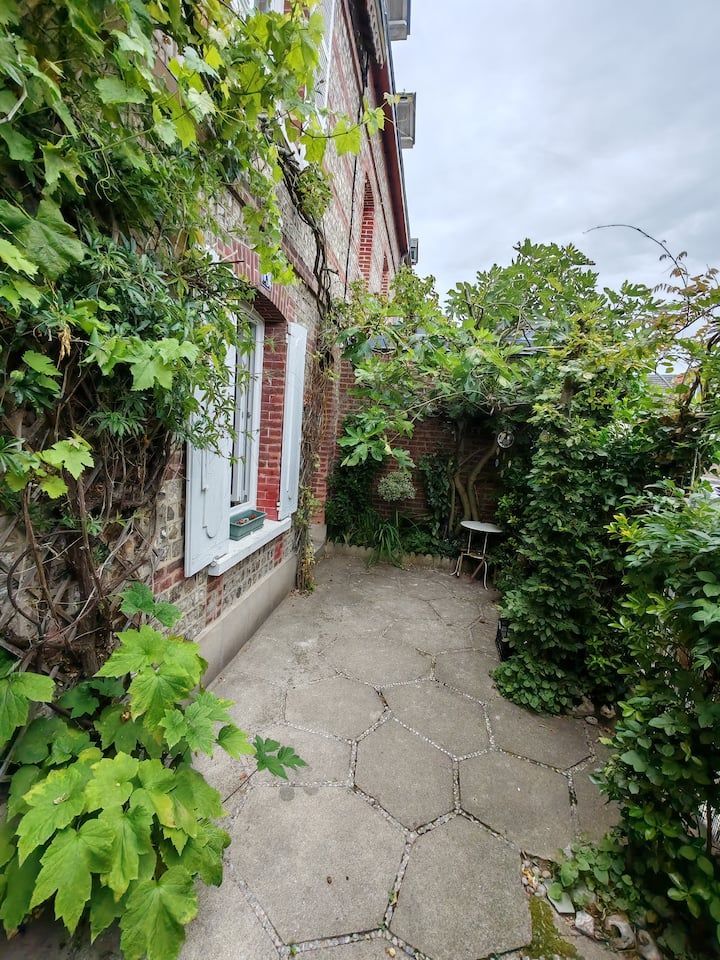 Terrasse en pierre avec une végétation luxuriante adossée à un bâtiment en briques doté d'une fenêtre et de volets blancs.