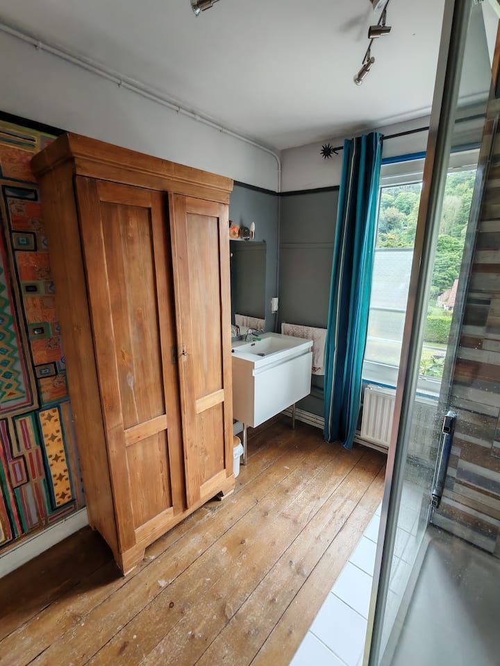 Armoire en bois dans une salle de bains avec lavabo, fenêtre et rideaux turquoise. Sol en bois.