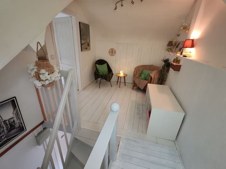 Vue du haut de l'escalier donnant sur un coin salon mansardé blanc avec deux fauteuils en osier, une petite table et une armoire blanche.