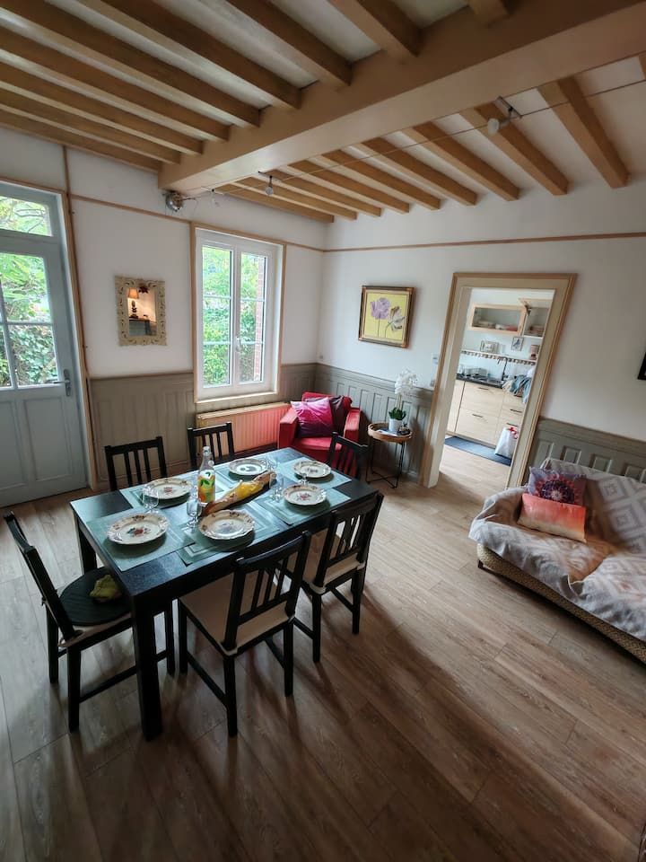 Salle à manger avec table dressée pour six personnes, fauteuil rouge, canapé et poutres apparentes.