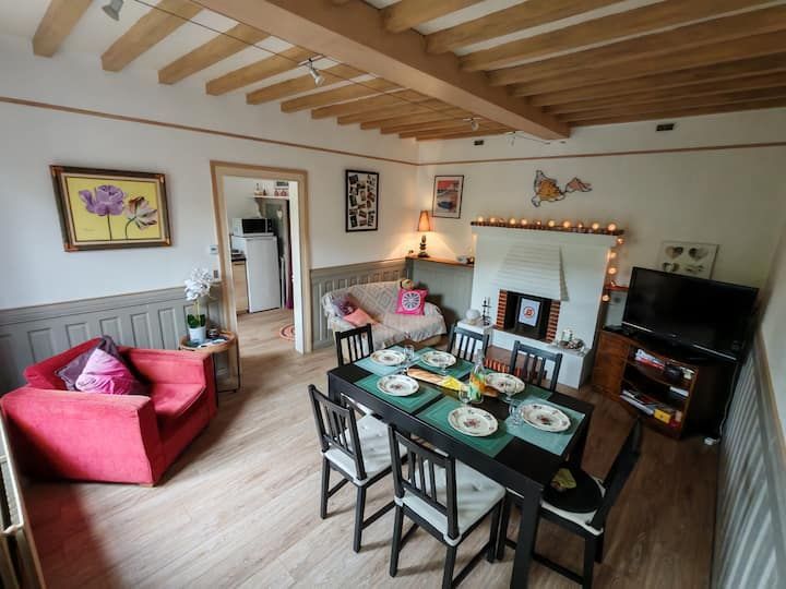 Coin repas et salon cosy : canapé rouge, table à manger pour six personnes, cheminée avec guirlandes lumineuses et vue sur la cuisine.