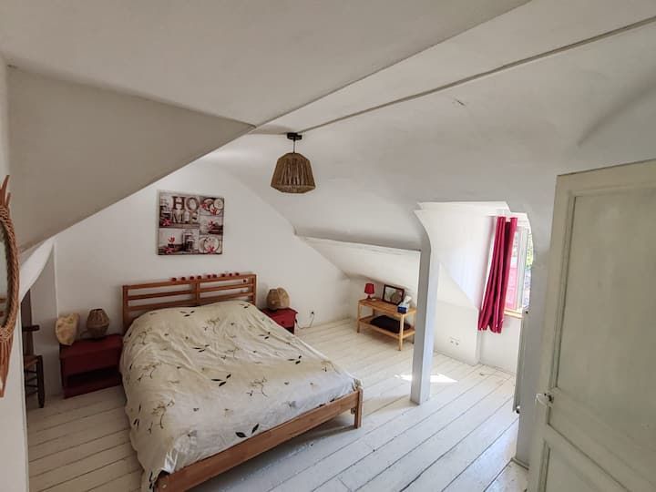 Chambre mansardée avec plafond en pente, lit en bois, murs et sol blancs et lumière naturelle.