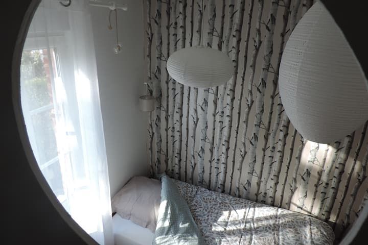 Coin de chambre avec un lit, des lampes blanches et du papier peint à motifs de bouleaux, vu à travers une ouverture circulaire.