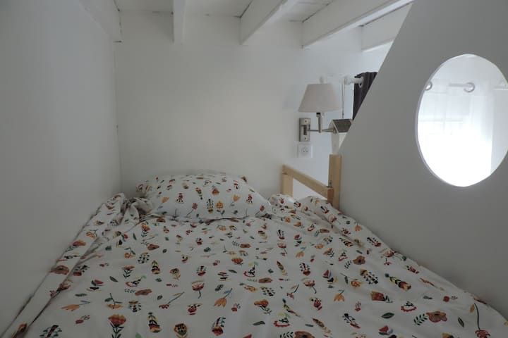 Chambre blanche étroite avec un lit. Linge de lit à motifs floraux, lampe de chevet et fenêtre ovale.