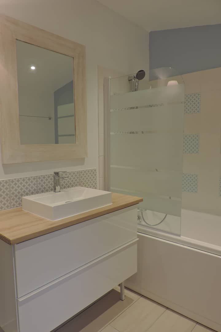 Salle de bains blanche avec lavabo, miroir et douche. Plan de travail en bois, touches de bleu et porte de douche en verre dépoli.