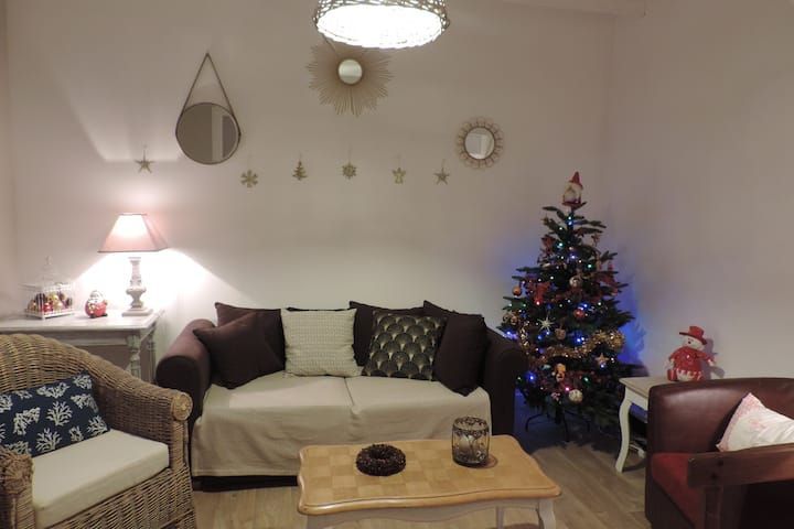 Salon chaleureux décoré pour Noël avec sapin, canapé, fauteuils et décorations murales.