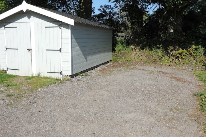 Un hangar gris jouxte une allée de gravier, avec de l'herbe à gauche et des arbres en arrière-plan.