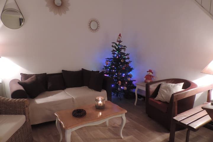 Salon chaleureux avec sapin de Noël. Canapé blanc et coussins marron, fauteuil rouge. Éclairage tamisé.