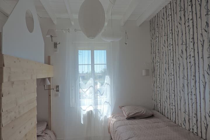 Chambre avec un lit superposé, un lit simple, des murs blancs, une fenêtre avec des voilages et du papier peint à motifs de bouleaux.