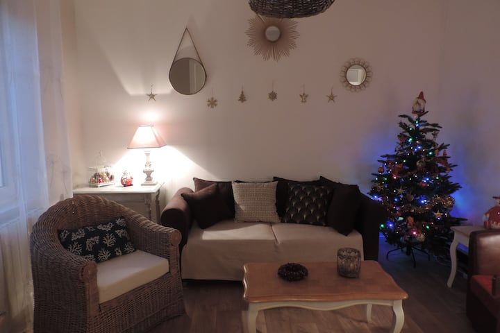 Salon chaleureux avec sapin de Noël, miroirs décorés, canapé et fauteuil en osier, éclairé par une lumière douce.