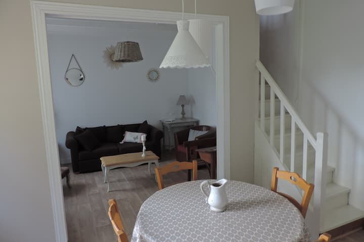 Coin repas avec table ronde pour deux, ouvert sur le salon avec canapé, fauteuil et escalier.