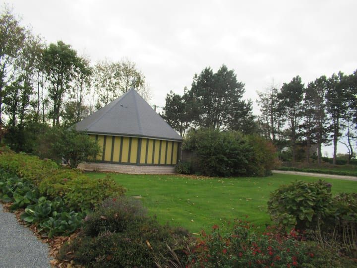 Petit bâtiment jaune et gris au toit pyramidal, entouré d'arbres et de buissons sur une pelouse.