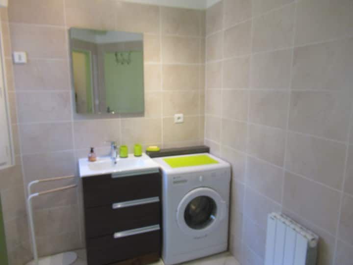 Salle de bains avec meuble-lavabo marron, machine à laver, miroir et murs carrelés beiges.