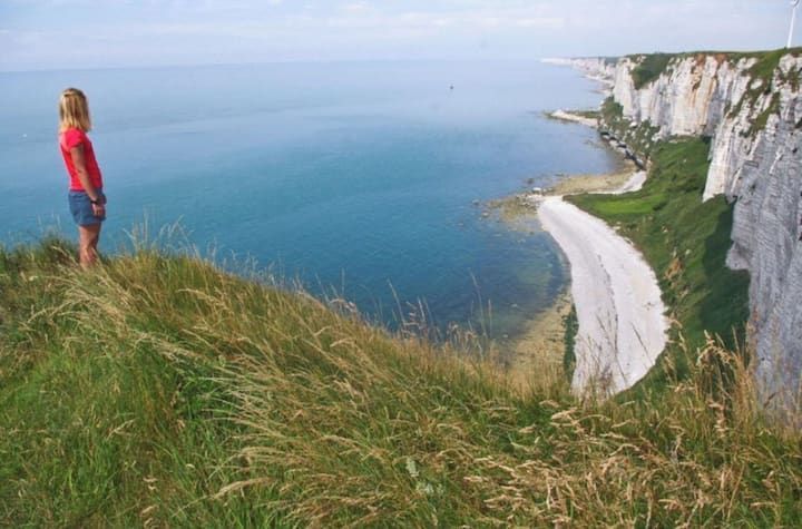 Femme contemplant l'océan du haut d'une falaise, falaises pâles, eau bleue et premier plan herbeux.